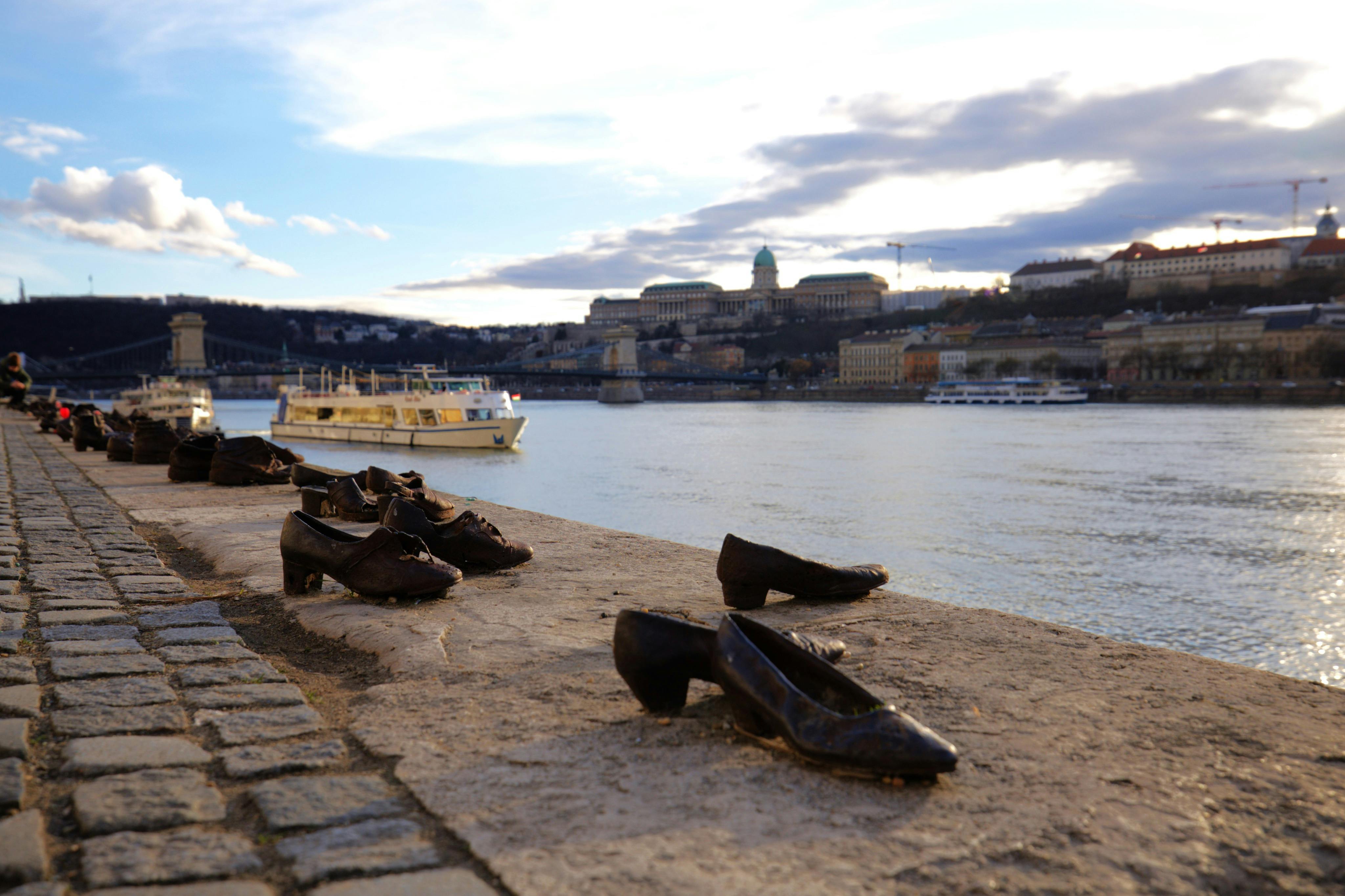Shoes on the Danube Bank memorial with a river, a boat, and historic buildings in the background under a partly cloudy sky.