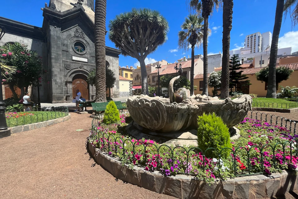 A garden with a stone fountain, flowers, and trees, adjacent to a dark stone building with an arched doorway under a blue sky.