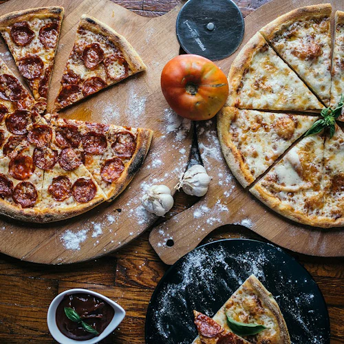 Två stora pizzor, en med pepperoni och en vanlig, på träbrädor med tomat, vitlök, pizzaskärare och sås.