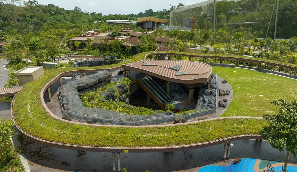 Aerial view of a park featuring a large modern building with a brown roof, surrounded by greenery, pathways, and nearby structures.