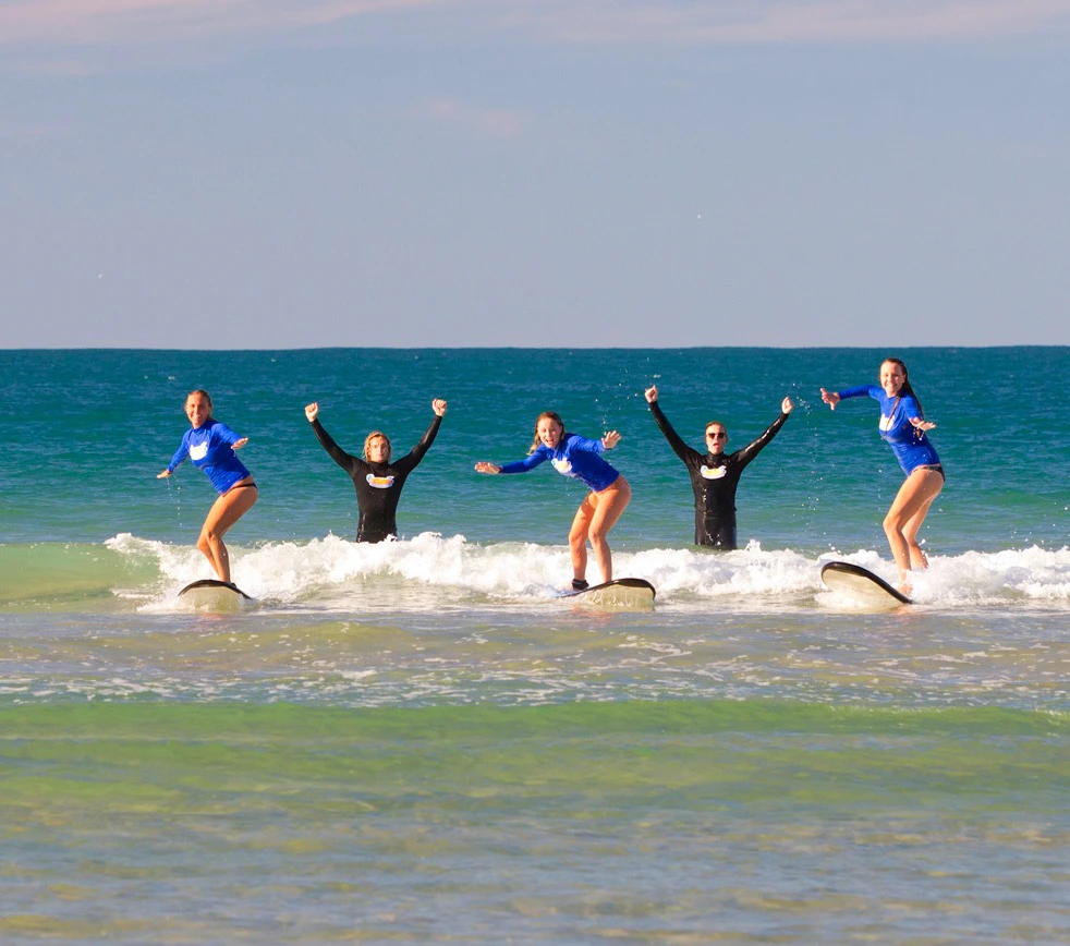 Five people in wetsuits surf in shallow ocean waves, raising their arms in apparent enthusiasm.