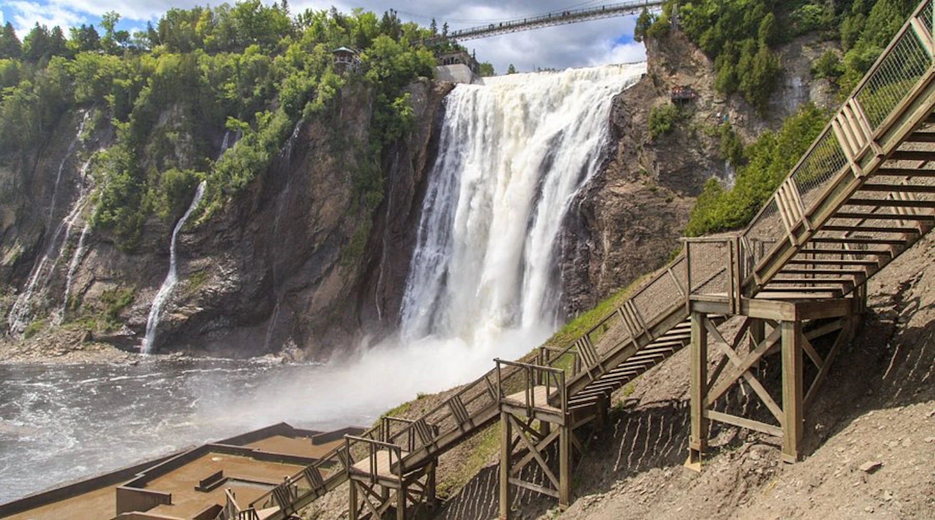 A powerful waterfall cascades down a rocky cliff, with a wooden staircase and viewing platform in the foreground. Trees surround the area.