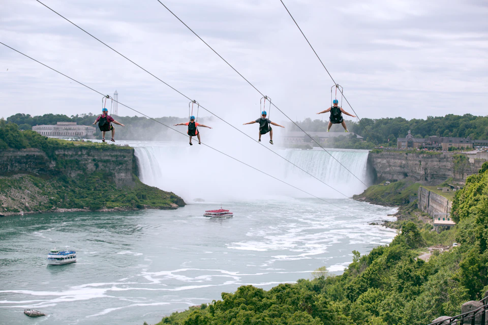Four people zip-lining in front of a large waterfall, with a boat on the river below and green foliage in the foreground.