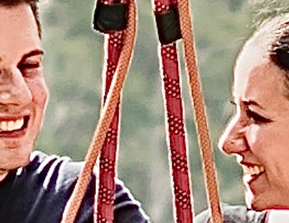 A man and woman are sitting on a swing suspended high above a lush, tropical forest with palm trees in the background.