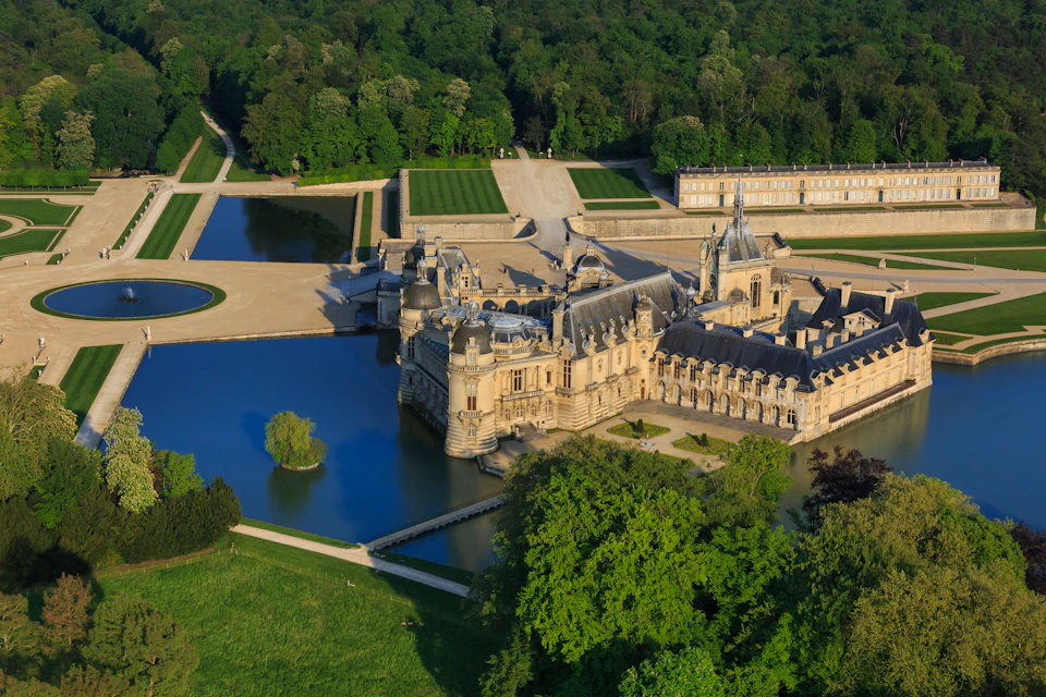 An aerial view of a large, historic castle surrounded by a moat, lush gardens, and dense forest.