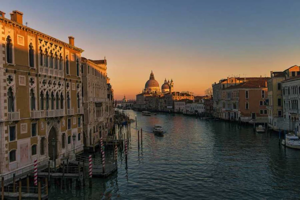 A canal in Venice at sunset, flanked by historic buildings, with a boat in the water and a dome in the background.