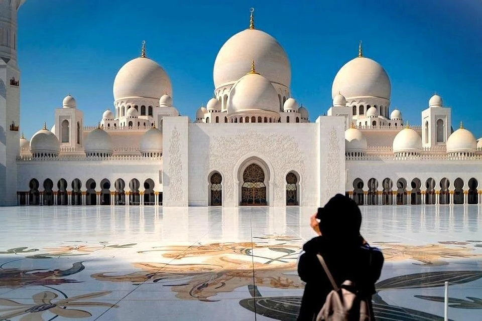 Person in black attire photographing a grand white mosque with large domes and intricate architecture under a clear blue sky.
