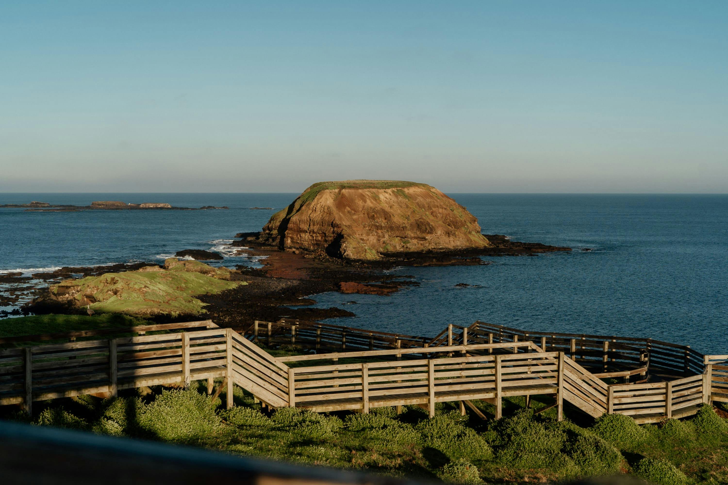 A rocky island with a grassy top in the ocean, viewed from a wooden boardwalk surrounded by greenery under a clear sky.