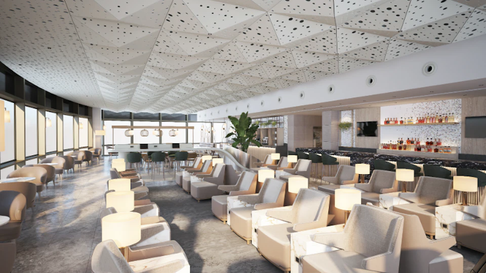 Modern lounge with numerous beige armchairs, small tables with lamps, a white geometric ceiling, bar area, and potted plants.