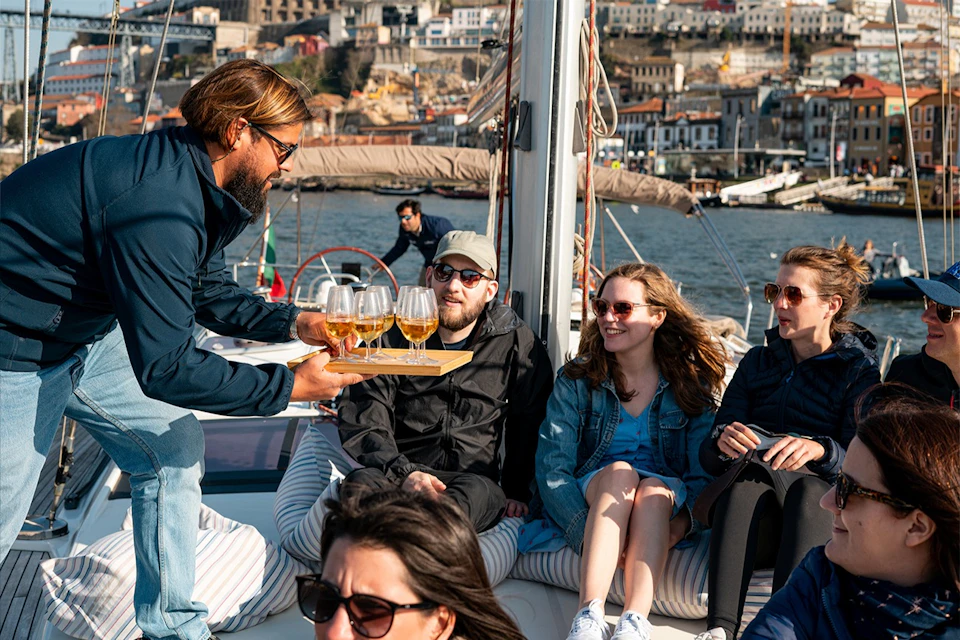 A group of people sitting on a boat, enjoying drinks served by a person. A cityscape is visible in the background.