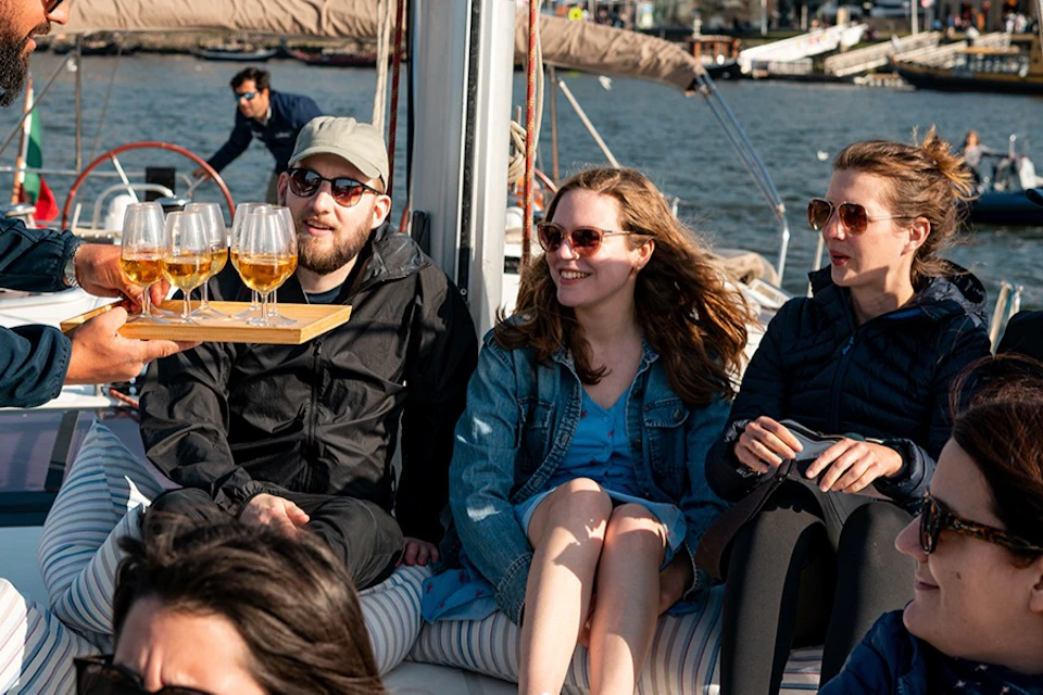 A group of people sitting on a boat, enjoying drinks served by a person. A cityscape is visible in the background.