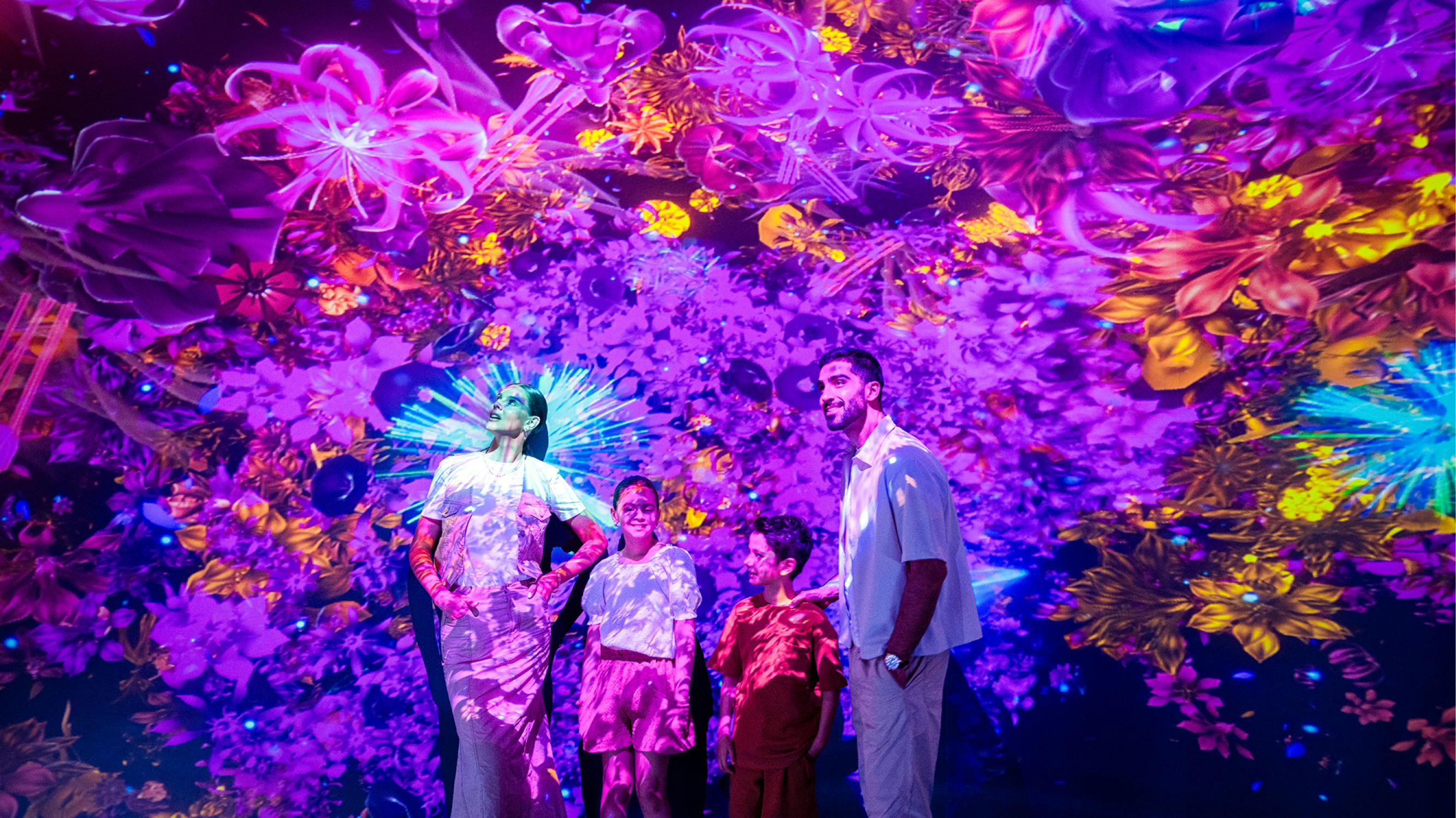 Τέσσερις άνθρωποι στέκονται μαζί, περιτριγυρισμένοι από ζωντανές, πολύχρωμες λουλουδένιες και αφηρημένες ψηφιακές προβολές στους τοίχους και την οροφή.