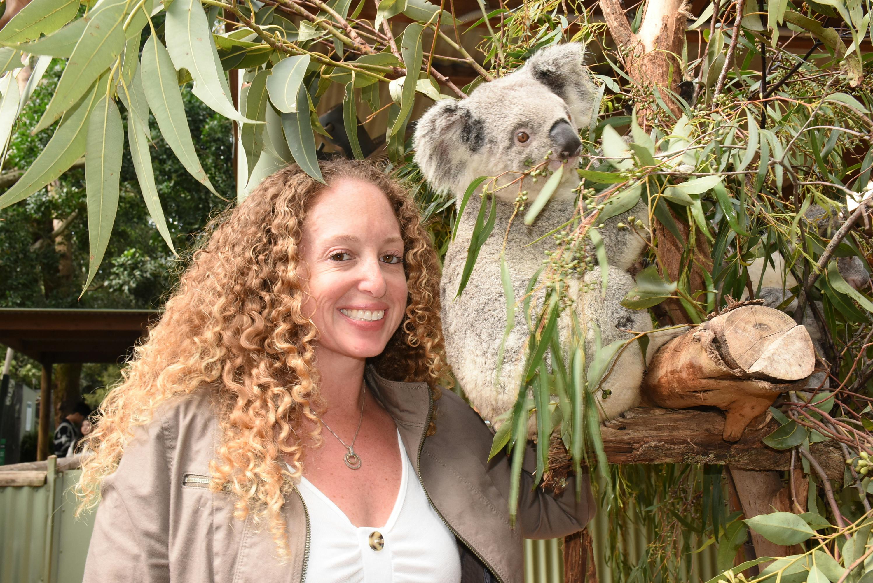 A person with curly hair smiles next to a koala perched on a tree branch surrounded by eucalyptus leaves.