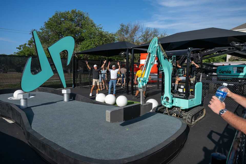 A group of people cheering near a mini excavator and large golf ball props on a platform under a clear sky.