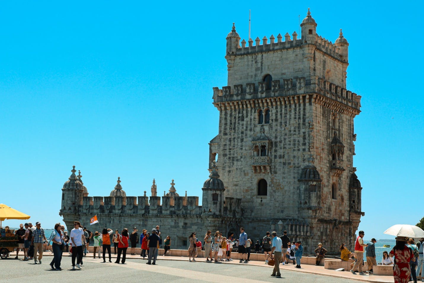 澄んだ青い空の下、城壁のある歴史的な石造りの建物、ベレンの塔の近くを歩く観光客。