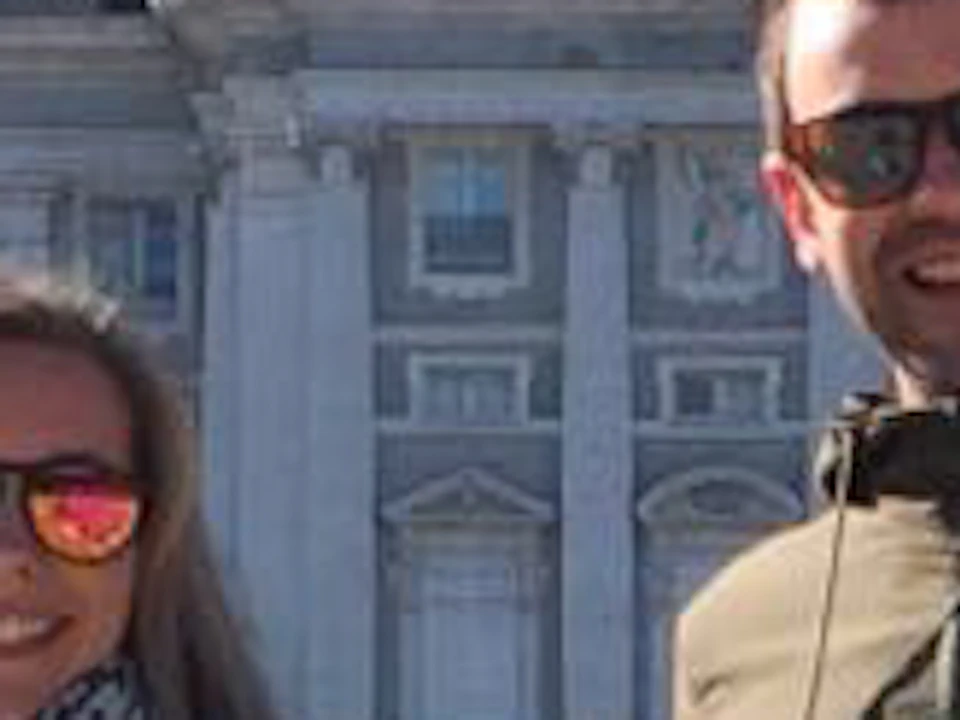 Two people standing with bicycles in front of a large, ornate building with a dome, surrounded by tall trees and under a clear blue sky.