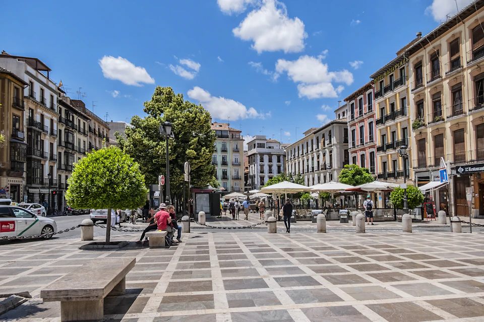 A sunny city square with trees, benches, people walking, and surrounding colorful buildings under a blue sky.