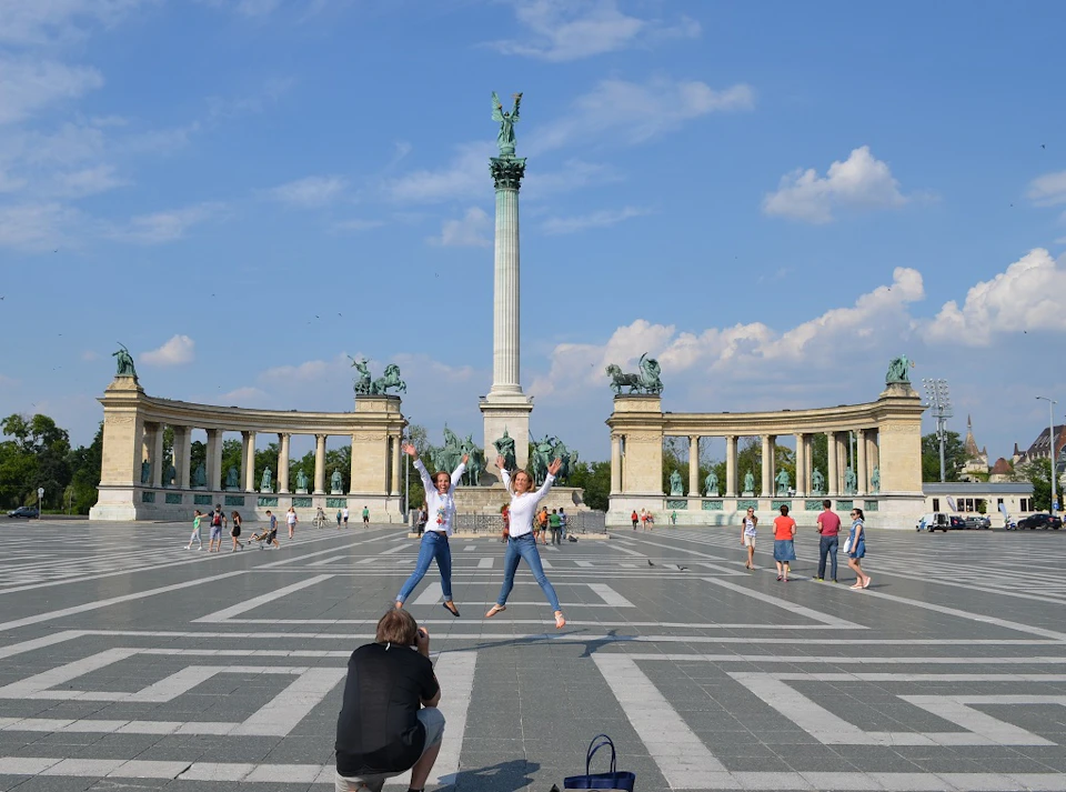 Two people jump mid-air while posing for a photo in front of a large monument with statues, with others walking around in the background.