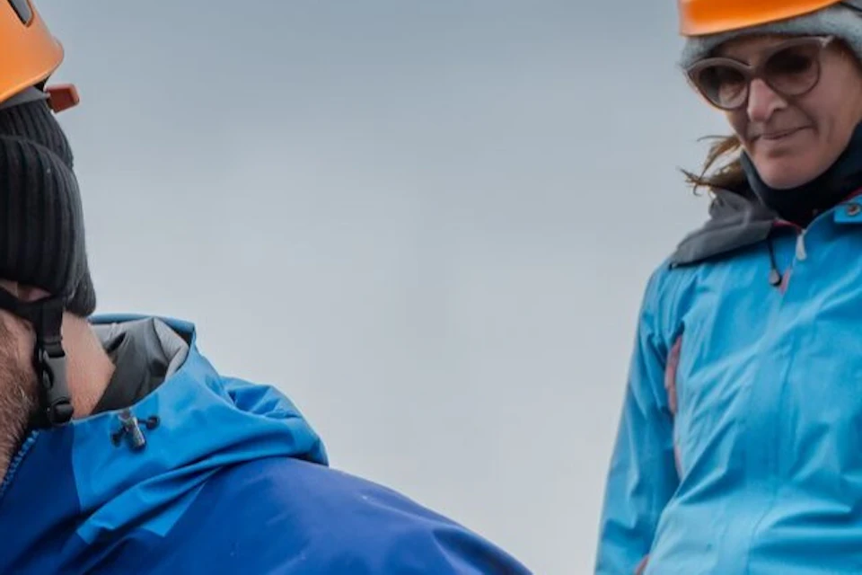 Three people in climbing gear, including orange helmets and blue jackets, walking on a snowy and rocky terrain under a cloudy sky.