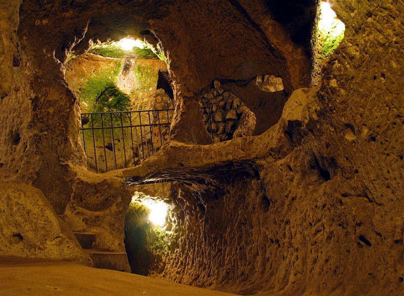 Grotta sotterranea illuminata con camere multiple, pareti ricoperte di muschio e una ringhiera di metallo.