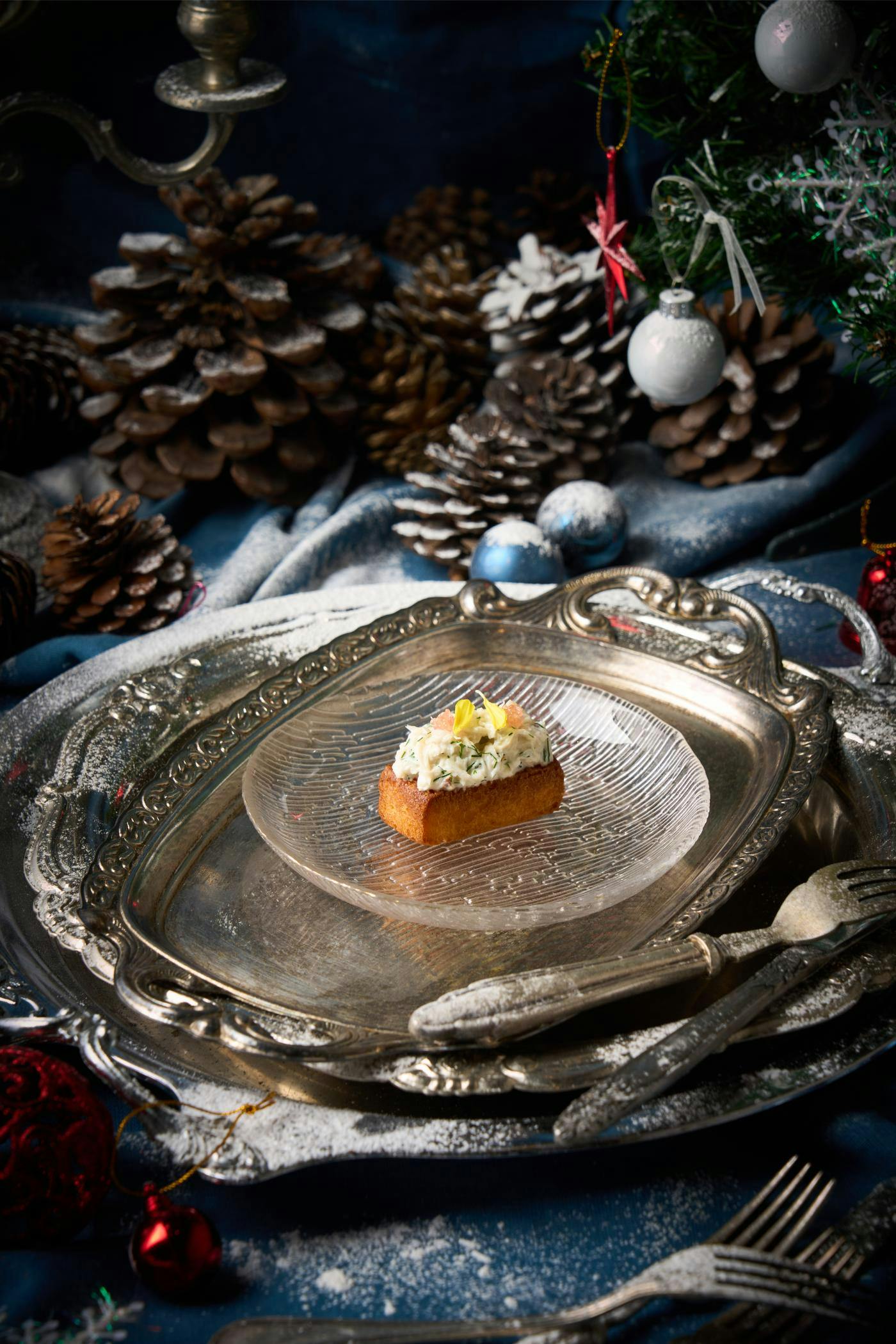 A piece of gourmet food on a clear glass plate, placed on a silver tray, surrounded by pinecones and festive decor.