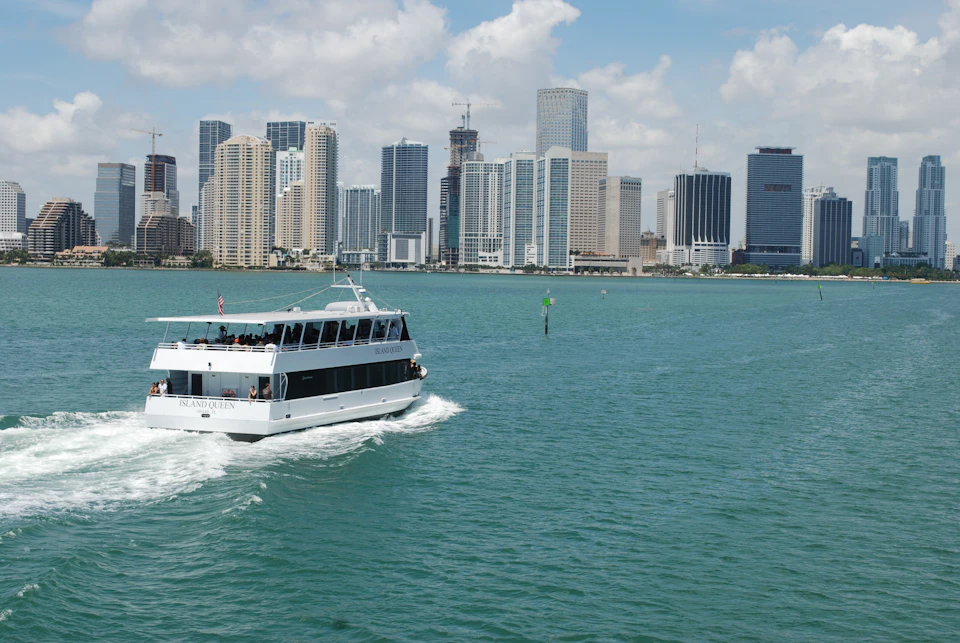 A white tour boat sails on green-blue water with a city skyline of tall buildings in the background under a partly cloudy sky.