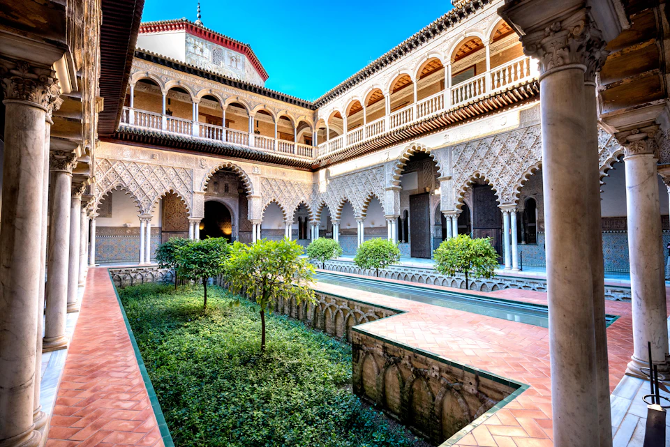 Historic courtyard with arched columns, intricate carvings, greenery, and a central water channel, framed by two-story balconies.