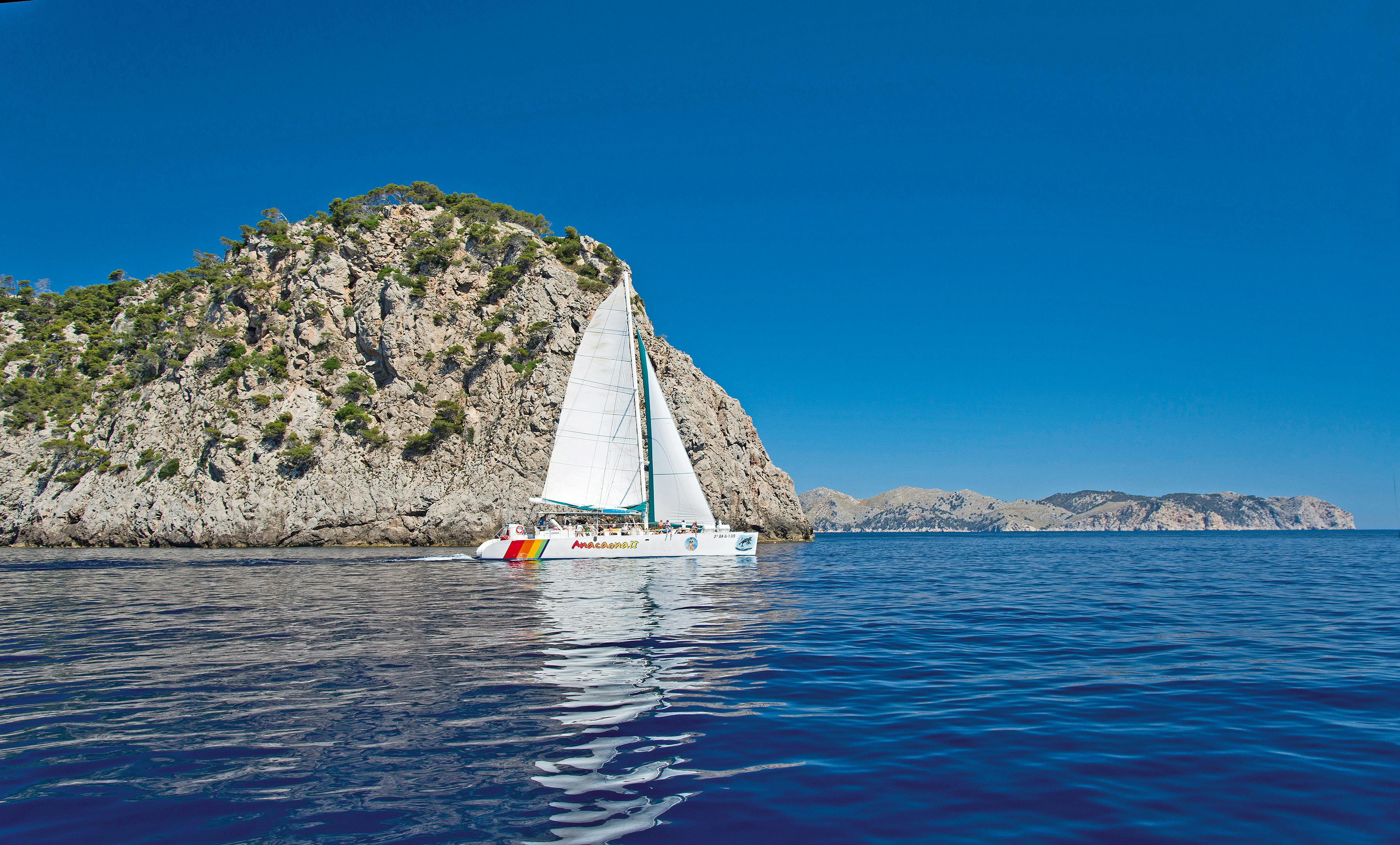 Un voilier blanc sur des eaux calmes et bleues, près d'une île rocheuse couverte de végétation, sous un ciel bleu clair.