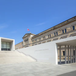 James-Simon-Galerie entré med breda trappsteg, modern arkitektur tillsammans med en klassisk byggnad under en klarblå himmel.