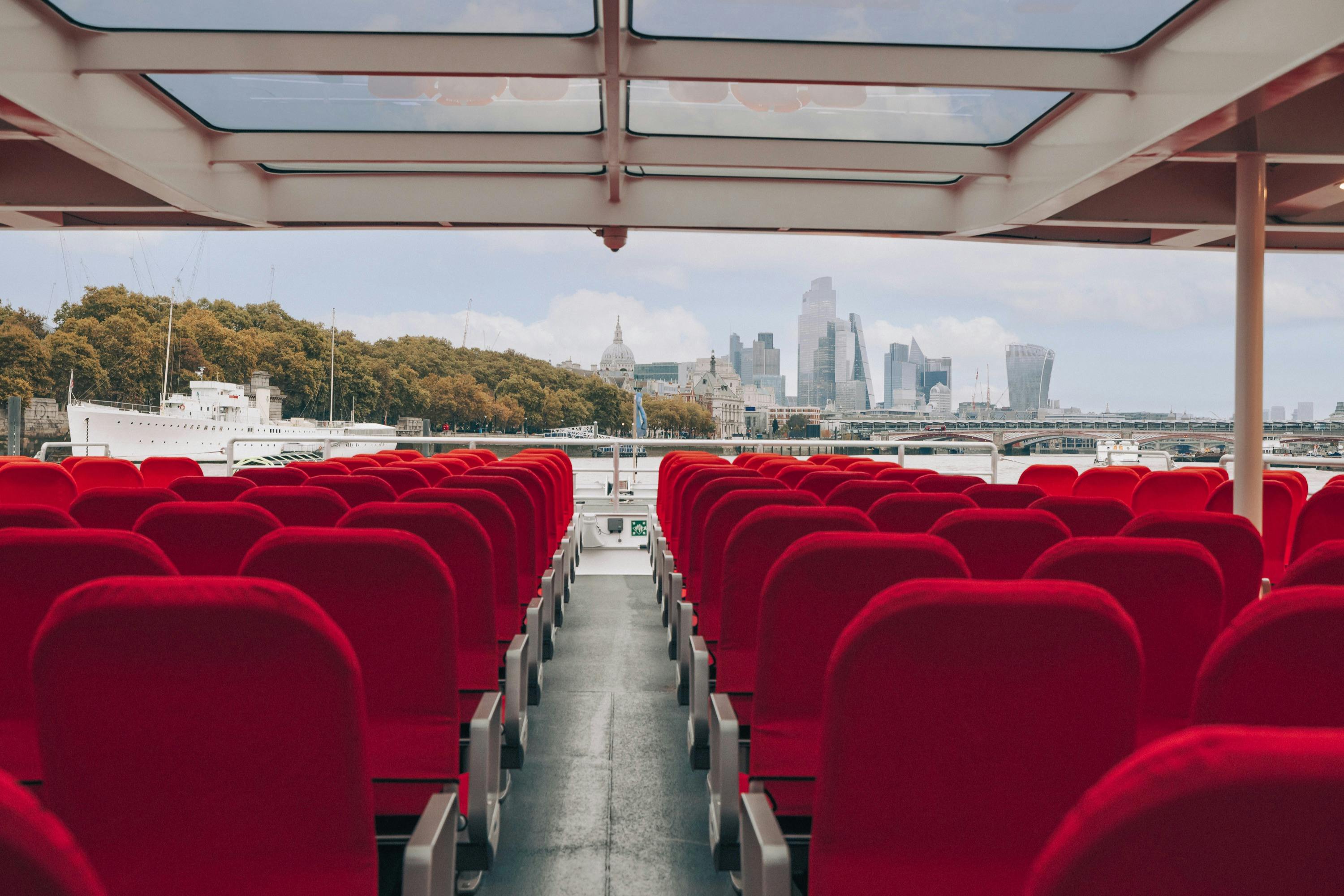 Rangées de sièges rouges vides sur un bateau avec un toit en verre, horizon de la ville et arbres visibles à l'arrière-plan.