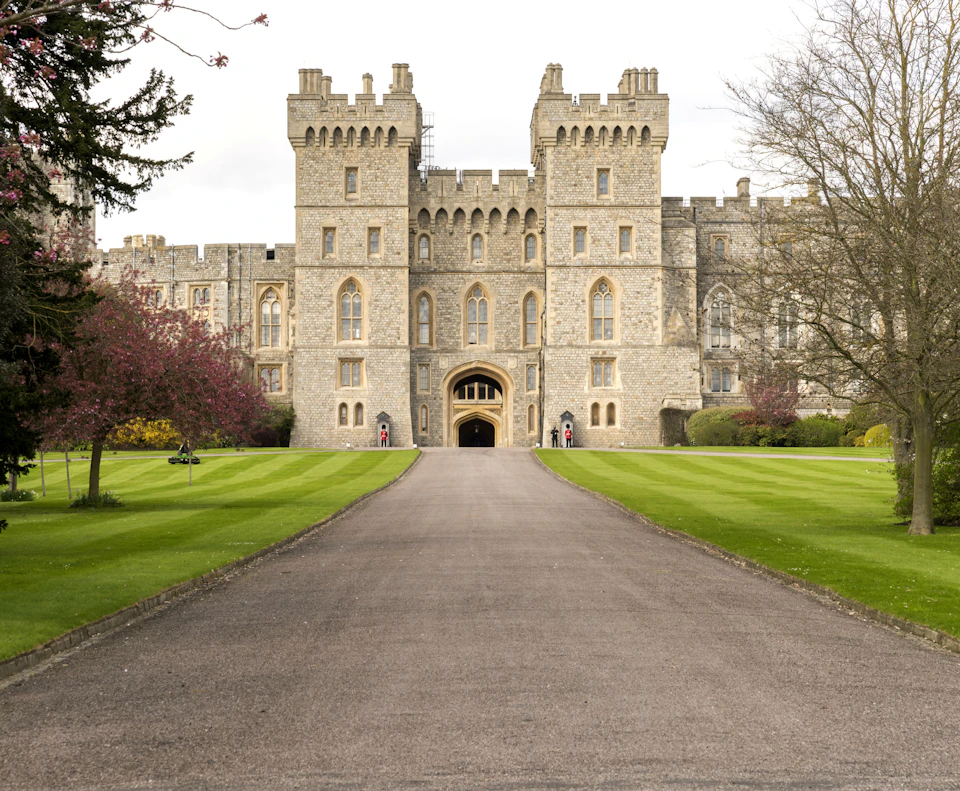 A large, historic stone castle with towers and battlements, surrounded by green lawns and trees, with a central pathway leading to the entrance.