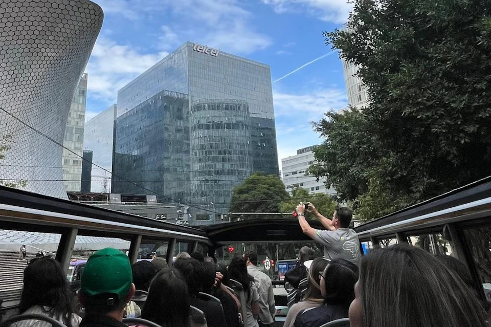 People on an open-top bus tour in a city with glass buildings, including one labeled "Telcel," and trees under a blue sky.