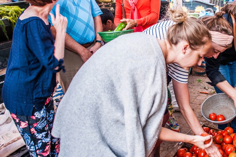 People shopping at an outdoor market, browsing and selecting produce from various stands, with one woman picking tomatoes.