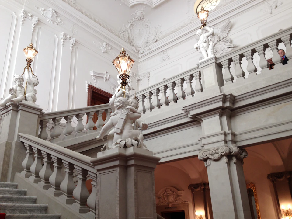 Elegant staircase with ornate railings, statues, and decorative lamps in a grand, white, intricately designed interior space.