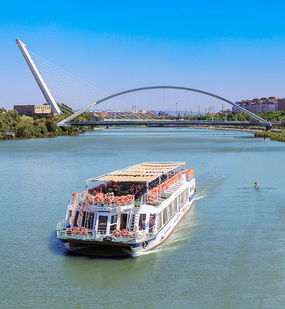 A white riverboat cruises under a modern arched bridge with cables, set against a backdrop of city buildings and a blue sky.