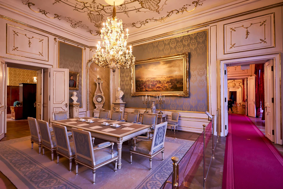 Lavish dining room with ornate chandelier, large framed painting, elegant table set for six, and intricate wall and ceiling designs.