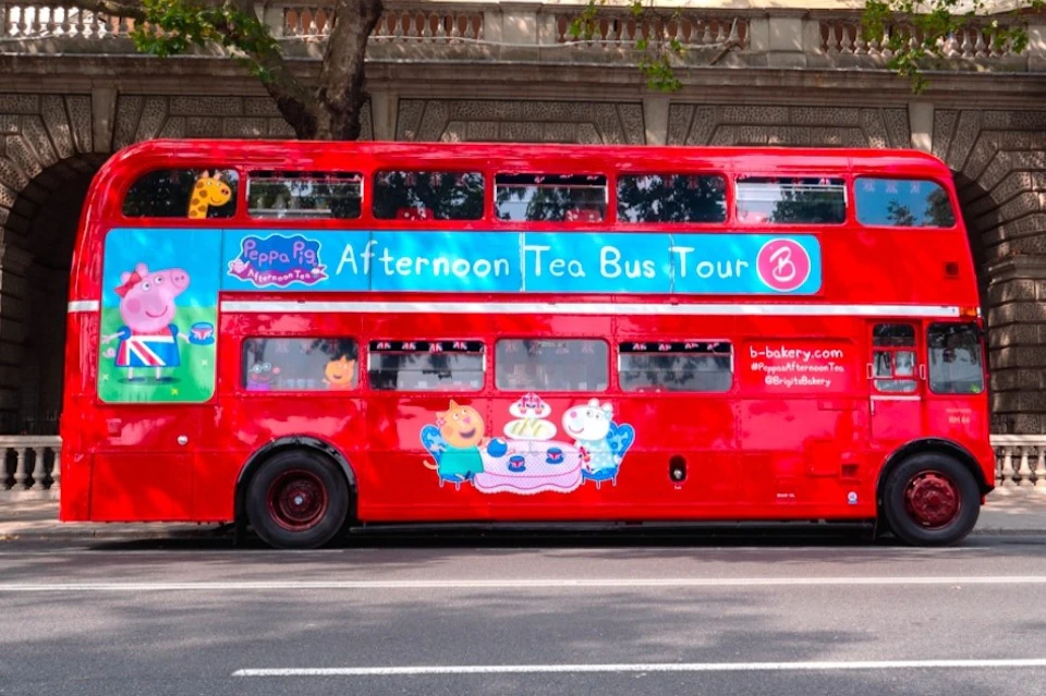 A red double-decker bus with Peppa Pig-themed decorations for an Afternoon Tea Bus Tour, parked on a street.