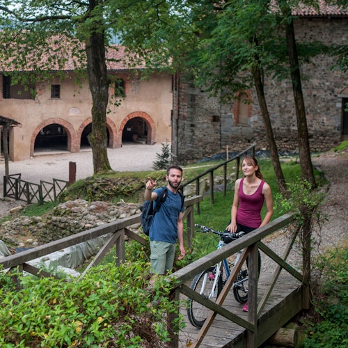 Due persone, una con una bicicletta, in piedi su un ponte di legno accanto a edifici storici in pietra e a una vegetazione lussureggiante.