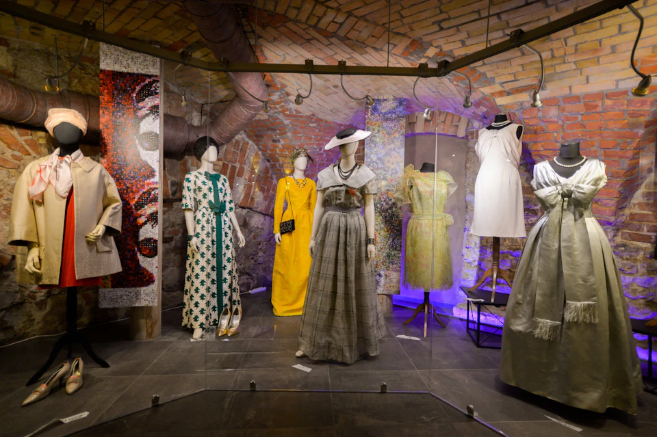 A display of vintage dresses on mannequins in a brick-walled room with spotlights highlighting the outfits.