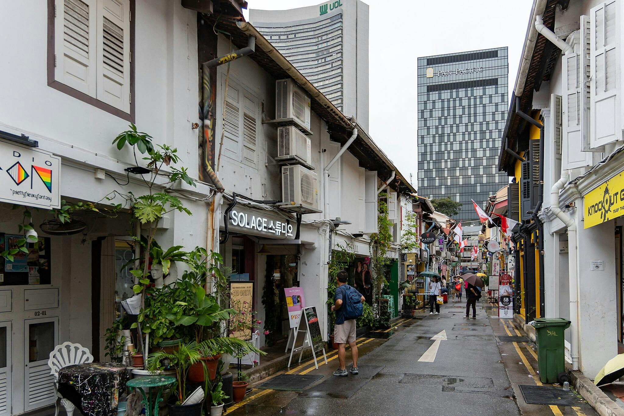 Pedestrian street with colorful shop fronts, signs, potted plants, and some people walking or browsing. Skyscrapers backdrop.