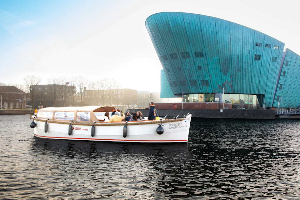 A boat with people on a canal near a modern, turquoise building under a partly cloudy sky.