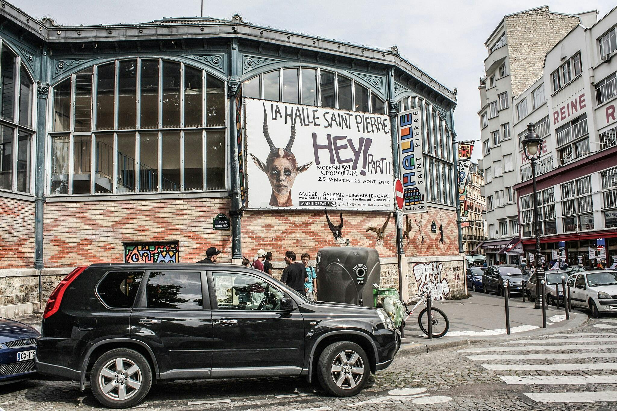 Внешний вид на La Halle Saint Pierre с большими окнами и рекламным щитом «ЭЙ! Современное искусство и поп-культура», припаркованные автомобили и пешеходы.