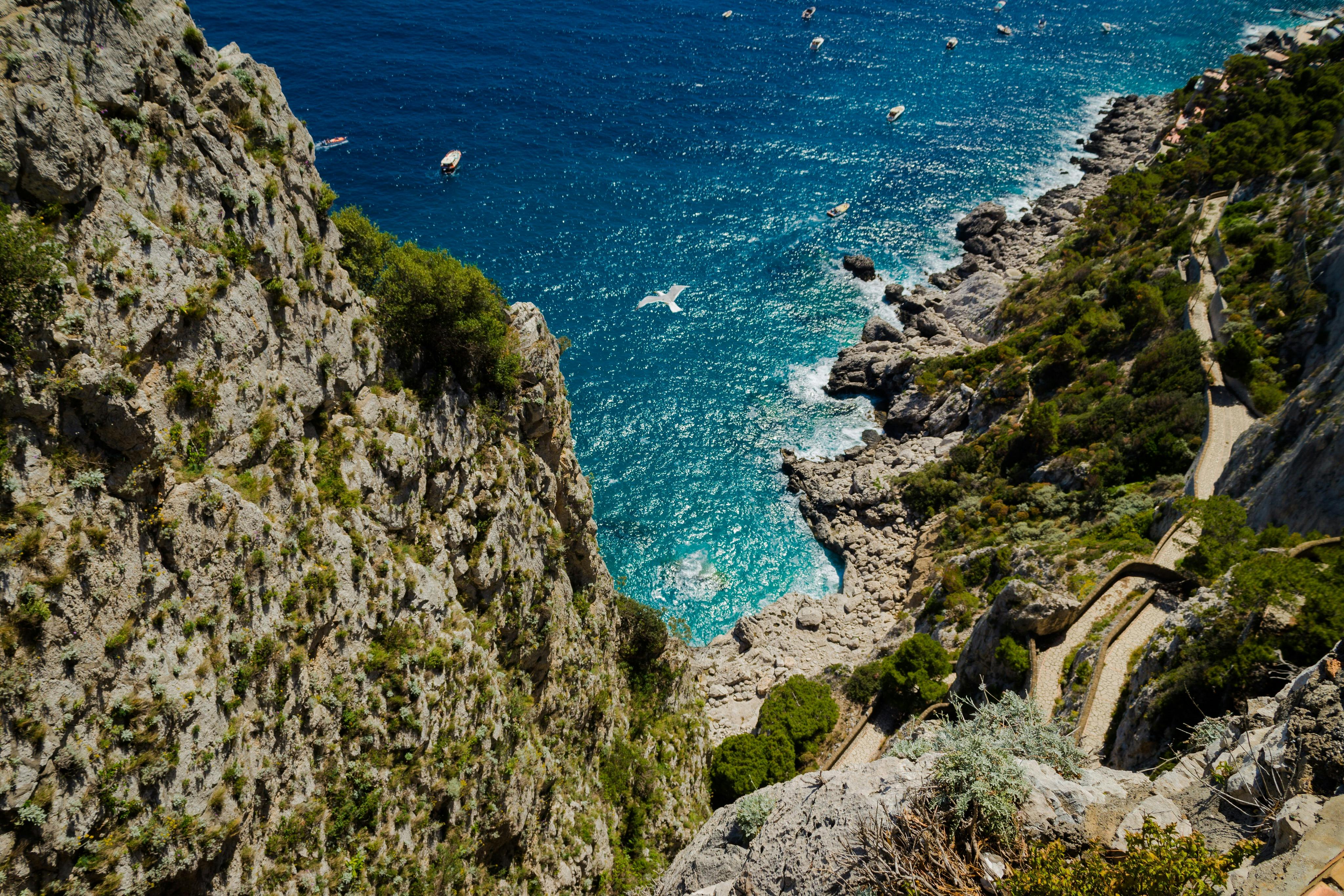 구불구불한 길이 펼쳐진 바위 해안의 공중 전망, 푸른 초목으로 뒤덮인 절벽, 푸른 바다 위의 보트.