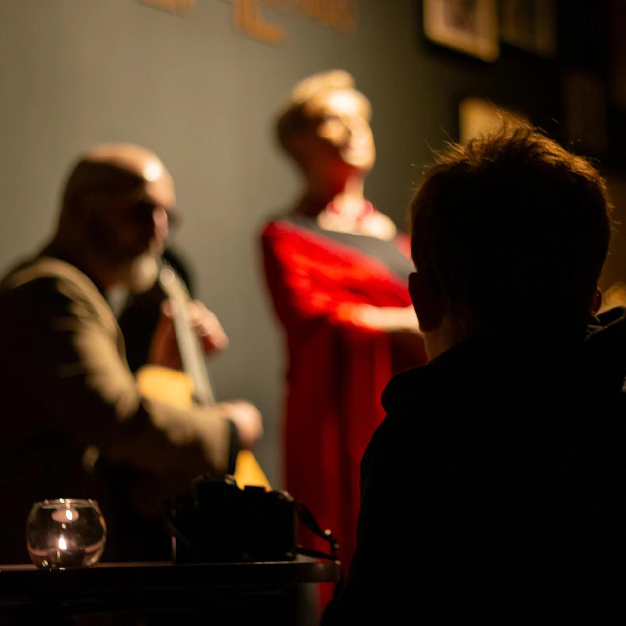The dimly lit scene shows a person watching a musician playing guitar and a singer performing, both slightly out of focus.