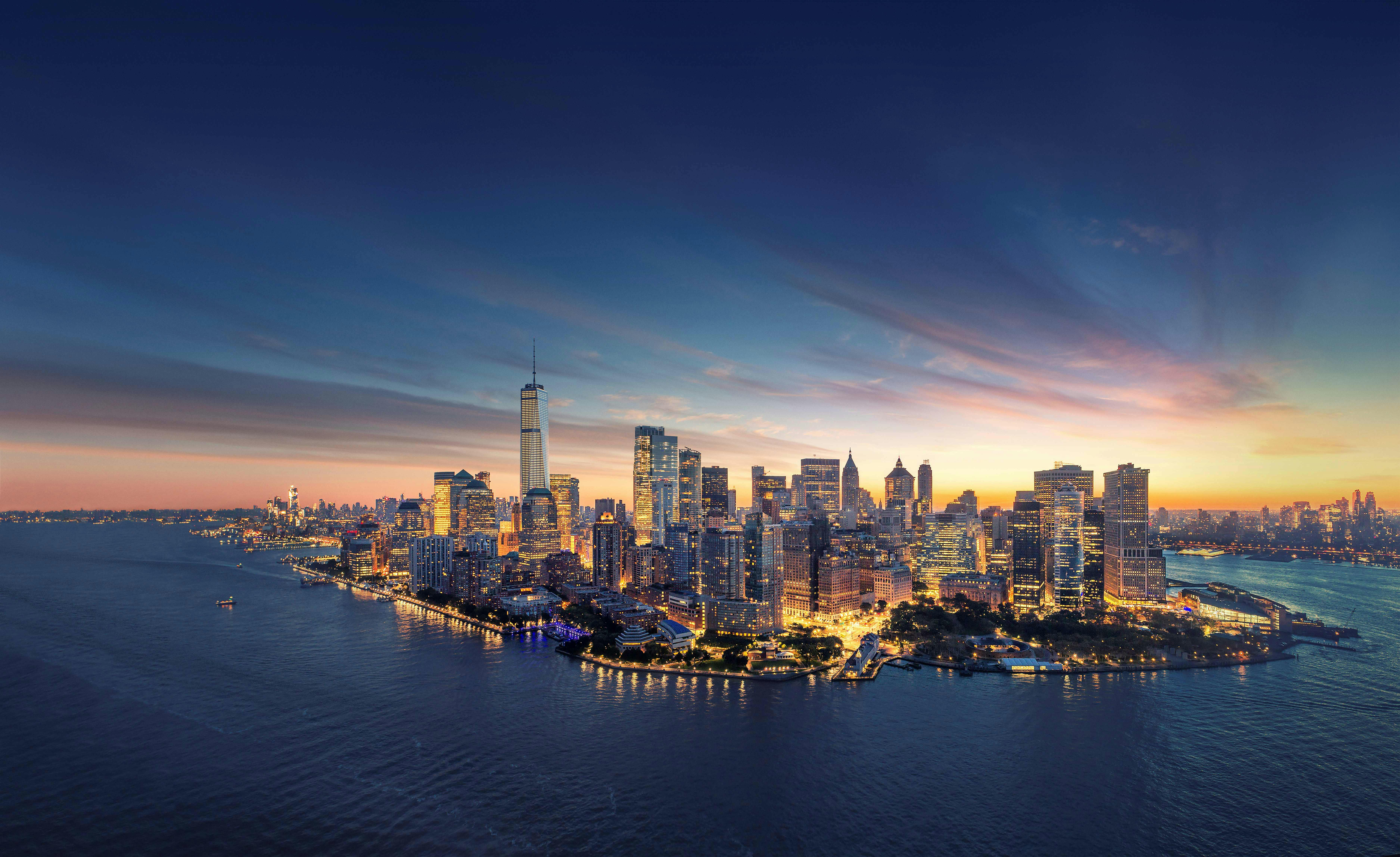 A city skyline at dusk with illuminated buildings along a waterfront, under a twilight sky.