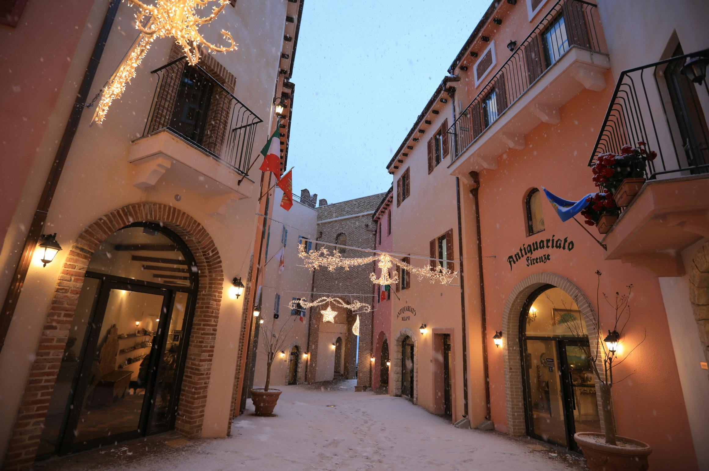 A narrow alley with buildings on both sides, lit lanterns, decorations, and snow on the ground under a dusky sky.