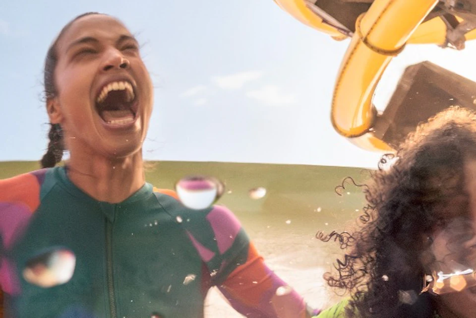 Three people laughing and enjoying a water ride with splashes, against a backdrop of a slide and sunlight.