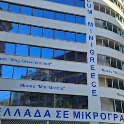 Museibyggnad med flera språk på fasaden, inklusive "Museum 'Mini-Greece'", och reflekterande glasfönster.