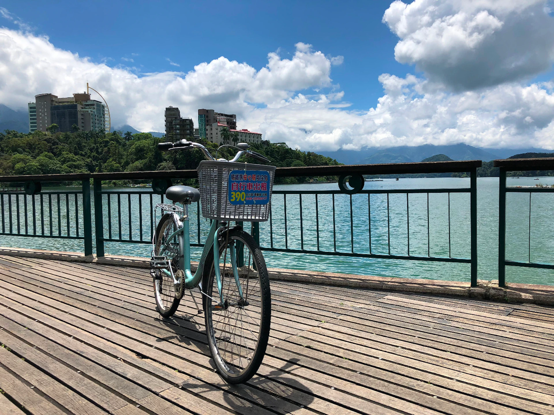 A teal bicycle with a basket stands on a wooden deck near a railing, overlooking a lake and distant buildings under a blue sky.