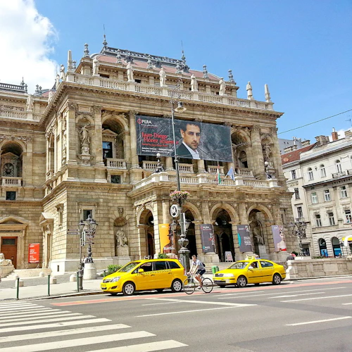 Stor historisk byggnad med utsmyckad arkitektur, människor som står vid en övergångsställe, gula taxibilar och en cyklist. Klar, blå himmel ovanför.